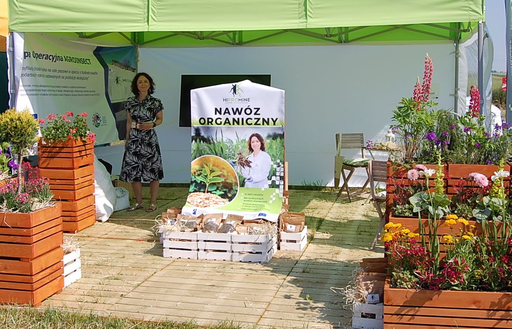 Zdjęcie przedstawia stoisko targowe z dużą ilością kwiatów i banerem na środku z napisem NAWÓZ ORGANICZNY. Przy banerze stoi kobieta prezentująca próbki firmy